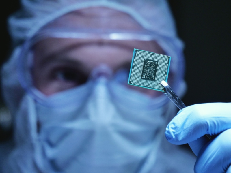 Cleanroom engineer holding a small chip with tweezers, wearing protective mask and gloves.